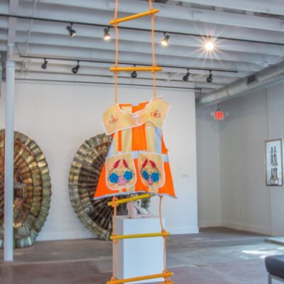 a decorated, high visibility workers vest hanging from a rope ladder. Titled Visible Labor by Angela Faz