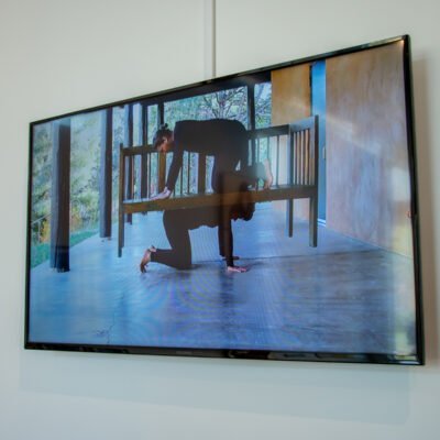 Still from Nida Bangash's video installation, The Bridge Called My Back. On a porch, one person on their hands and knees holds a wooden bench on their back as another person kneels on top on their hands and knees