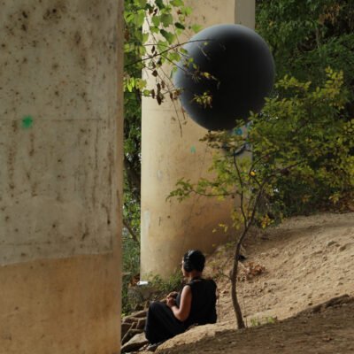 Production still of Ariel René Jackson sitting by pillars holding black balloon, photographed by Hiram Mojica