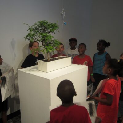 Children smiling at a sculpture that waters a plant.