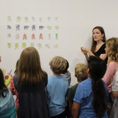 Female art educator speaking to children in front of art on the wall.